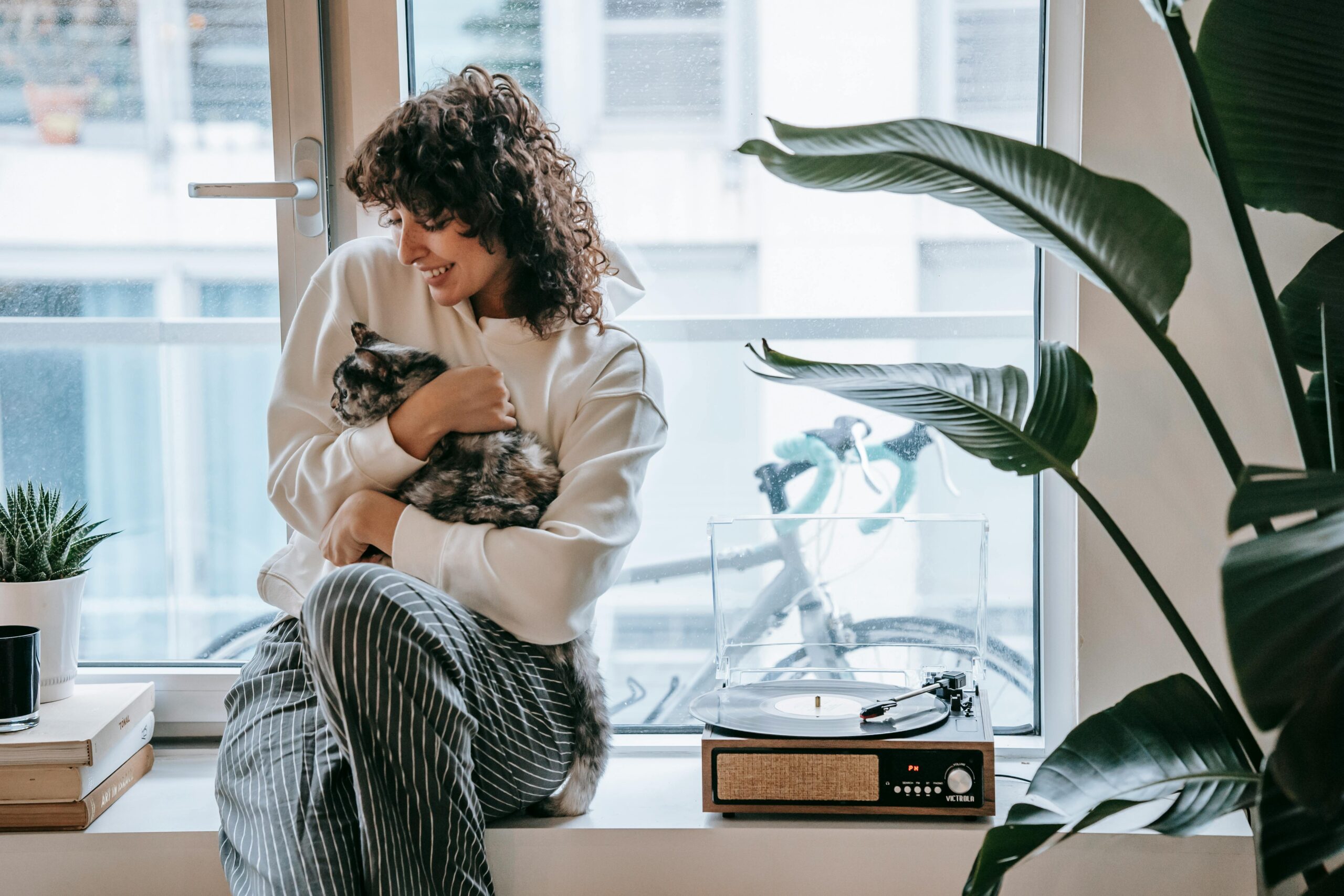 Young woman in cozy attire hugging her cat by the window, surrounded by plants and a retro turntable.